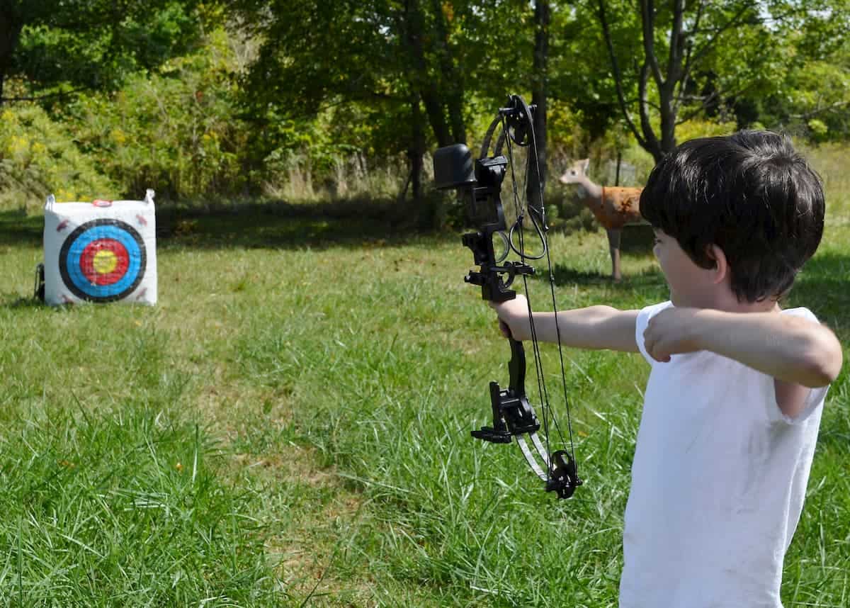 A dark haired boy holds his bow out towards the target as he practices archery on a sunny day - What is the Best Age to Start Archery
